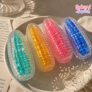 May include: Four oval-shaped, textured squishy toys with a clear outer shell and a vibrant inner core in teal, yellow, pink, and blue. The toys feature a grid-like pattern and are labeled with the text Sakura Squish in the upper right corner.