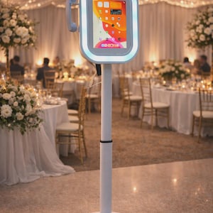 May include: A white photo booth with a touch screen display, a telephone handset, and a circular base. The screen displays a colorful interface. The booth is set up in a wedding reception hall with tables and floral arrangements.