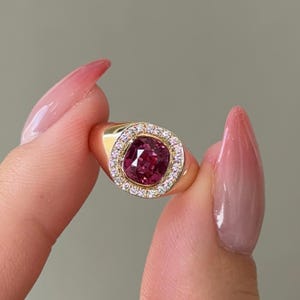 May include: A gold ring with a large, square-cut, deep pink gemstone encircled by a halo of small, clear diamonds. The ring's band is plain, polished gold. The ring is held up by a person's fingers.