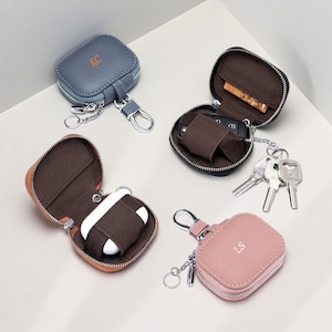 May include: Four small, square, zippered cases with rounded corners and metal key ring attachments. Two cases are closed, one blue with 'KC' and one pink with 'LS' monogrammed. Two cases are open, one black holding keys and one orange holding earbuds.