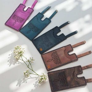 May include: Five rectangular leather luggage tags in purple, teal, dark blue, brown, and dark brown. Each tag features a cutout top, a strap with a metal snap, and a stamped geometric line design. Two tags are personalized with the initials GM and LK.