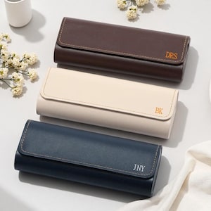 Peut inclure: Trois étuis à lunettes rectangulaires avec des rabats et des coutures visibles. L'étui du haut est marron foncé avec les lettres dorées DRS. L'étui du milieu est crème avec les lettres dorées BK. L'étui du bas est bleu marine avec les lettres argentées JNY.