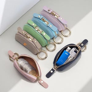 May include: A collection of six small, textured, zipped pouches with attached gold-toned key rings. The pouches come in lavender, light blue, green, beige with the letters MC, pink, and navy blue. One pink pouch holds silver coins, and the navy pouch holds a blue USB drive and keys.