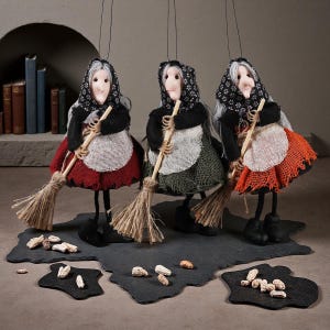 Peut inclure: Trois figurines de sorcières suspendues avec des cheveux gris, des foulards noirs à motifs floraux blancs, des hauts noirs et des tabliers blancs. Chaque figurine porte une jupe rouge, verte ou orange, tient un balai et a des chaussures noires.