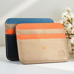May include: Two rectangular leather card holders with rounded corners and visible stitching. The front holder is beige with an orange stripe and the initials LK embossed. The back holder features blue, beige, orange, and dark navy horizontal panels.