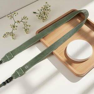 May include: A sage green camera strap featuring a textured surface, white contrast stitching, and the letters KK embossed on one end. The strap includes black plastic hardware connectors at both ends for attachment to a camera body.