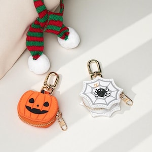 May include: Two small zippered pouches with gold-tone metal clasps. One pouch is orange and shaped like a jack-o'-lantern with a black face. The other is white, shaped like a spiderweb, and features a black spider and the letters DU.