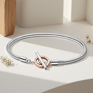 Puede incluir: Una pulsera plateada con un diseño de cadena de serpiente. La pulsera presenta un cierre de palanca con un acento de oro rosa y plata. La pulsera se exhibe sobre una superficie blanca con iluminación suave.