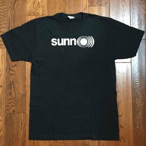 May include: A black short-sleeved t-shirt featuring the white text "sunn" followed by a circular graphic with concentric rings on the chest. The garment has a crew neckline and a standard fit, displaying a clean and simple design on the front.