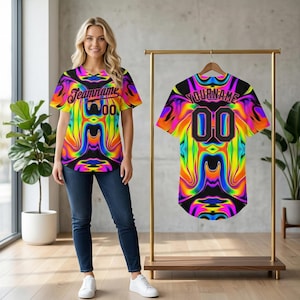 Puede incluir: Una vibrante camiseta de béisbol colorida con un diseño psicodélico arremolinado. La camiseta presenta el texto "Teamname" y el número "00". La camiseta se muestra en una percha dorada.
