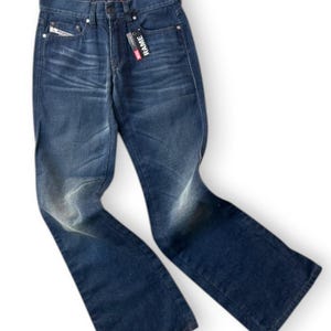 May include: Dark blue denim jeans featuring a faded wash effect on the thighs and knees. The jeans have a classic five-pocket design with a button fly closure and a black brand tag attached to the front pocket that reads RAME in white letters.