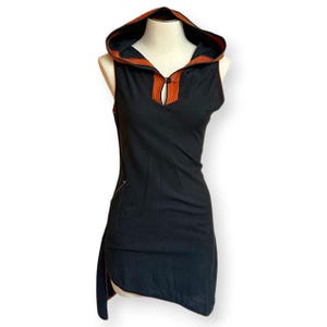 May include: A sleeveless black tunic featuring a hood with a burnt orange interior lining. The garment includes a deep V-neckline with a single button closure, a side pocket, and an asymmetrical hemline that is longer on one side.