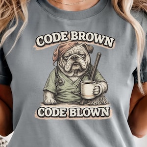 以下が含まれることがあります： 医療をテーマにしたブルドッグのイラストが描かれたグレーのTシャツ。ブルドッグは帽子とスクラブを着て、コーヒーマグと医療器具を持っています。シャツのテキストは、イラストの上に「CODE BROWN」、下に「CODE BLOWN」と書かれています。