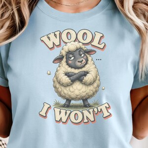 May include: Light blue t-shirt featuring a cartoon sheep with crossed arms and the text "WOOL I WON'T". The sheep is depicted with fluffy, cream-colored wool and a dark gray face. The shirt has rolled-up sleeves.