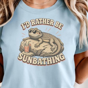 May include: Light blue t-shirt with the text "I'D RATHER BE SUNBATHING" and a graphic of a lizard wearing sunglasses on a rock. The shirt has rolled-up sleeves and a crew neck.