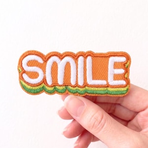 May include: An orange embroidered patch with the word "SMILE" in white block letters. The patch has a green and yellow border and is held by a hand.
