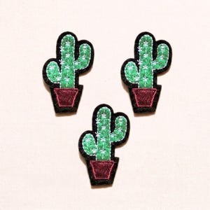 May include: Three embroidered cactus patches. Each patch features a green cactus with white accents in a maroon pot, outlined in black. The patches are likely for clothing or accessories.
