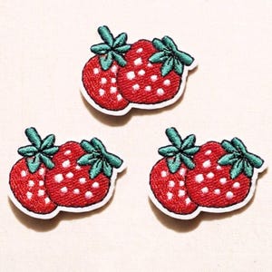May include: Embroidered patches featuring a pair of red strawberries with white seeds and green leaves. The patches have a white border and are arranged on a light background. These patches are ideal for adding to clothing or accessories.