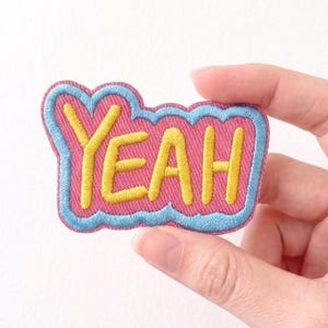 May include: Embroidered patch with the word "YEAH" in yellow, outlined in blue, on a pink background. The patch has a cloud-like shape and is designed for application on clothing or accessories.