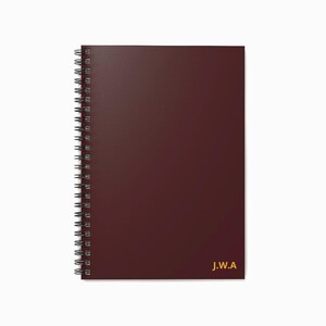 Puede incluir: Un cuaderno de espiral burdeos con una cubierta lisa. El cuaderno tiene una encuadernación en espiral plateada y letras doradas que dicen "J.W.A" en la esquina inferior derecha. El cuaderno es rectangular y tiene un diseño limpio.