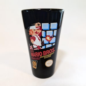 May include: Black shot glass featuring a Super Mario Bros. design. The glass showcases Mario in a pixelated style, with the game's logo and Nintendo branding. A collectible item for video game enthusiasts.