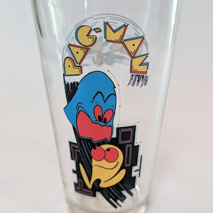 Könnte beinhalten: Klares Glas mit einem Pac-Man-Design. Das Glas zeigt einen blauen Geistercharakter mit roten Augen und einen gelben Geistercharakter mit roten Augen. Das Pac-Man-Logo ist in Gelb, Rot und Blau.