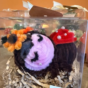 May include: A clear box containing two decorative floral arrangements. One is a brown and pink flower with pearl accents, and the other is a red flower with pearl accents. The box includes shredded paper and a label that reads "Petite Bloom Box."
