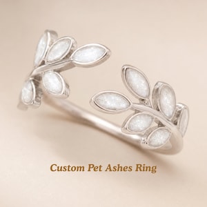 May include: A silver-toned open ring featuring two mirrored leaf-shaped branches. Each leaf contains a textured white inlay. The text Custom Pet Ashes Ring is printed in brown at the bottom of the image. The ring has a polished metallic finish.