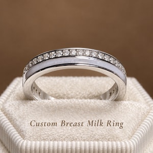 May include: A silver-toned ring featuring a row of small, clear, sparkling stones along the top edge and a smooth, opaque, off-white inlay band below. The text "Custom Breast Milk Ring" is printed in a dark font below the ring.