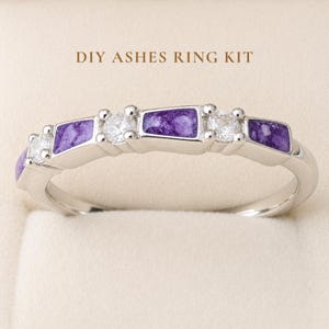 May include: A silver-toned ring featuring alternating purple stone inlays and clear round faceted stones. The text DIY ASHES RING KIT is printed above the ring. The band has a polished metallic finish and a curved design with multiple settings.
