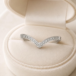 May include: A silver-toned chevron-shaped ring featuring a row of small, clear, round-cut stones embedded along the top surface. The ring is displayed inside a cream-colored, velvet-lined jewelry box with a rounded, hinged lid design.