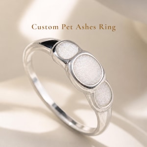 May include: A silver-toned ring featuring three circular settings, each containing a textured white inlay. The text Custom Pet Ashes Ring is displayed above the jewelry piece, which has a polished metal band and a smooth, reflective finish.