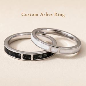 May include: Two metal rings with the text Custom Ashes Ring above them. One ring features a black textured inlay divided into segments, while the other ring features a smooth white inlay divided into segments. Both rings have a polished finish.