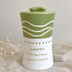 May include: A cylindrical ceramic urn with a lid, featuring a sage green top section with carved wavy lines and a white bottom section. The white area displays the name Lauren, the text In My Heart Always, and the dates 9/14/2017-2/16/2026 in green.