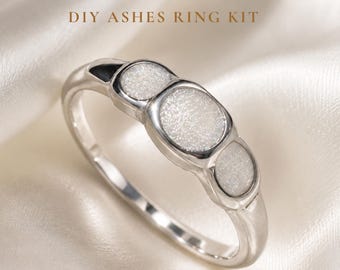 May include: A silver-toned ring featuring three circular settings filled with a textured, shimmering white material. The text DIY ASHES RING KIT is printed in brown above the ring. The band is smooth and polished, showcasing a clean, metallic finish.