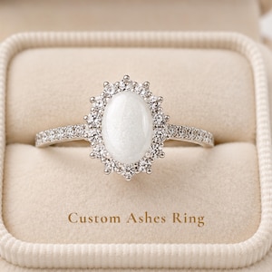 May include: A silver-toned ring featuring a central oval-shaped white stone surrounded by a halo of small clear stones. The band is also set with small clear stones. The text Custom Ashes Ring is printed below the ring on the beige velvet surface.