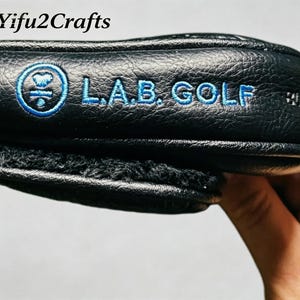 May include: Black golf club head cover with blue embroidered text and logo. The text reads "L.A.B. GOLF". The cover has a textured surface and a soft, fuzzy interior lining. The brand name "Yifu2Crafts" is in the top left corner.