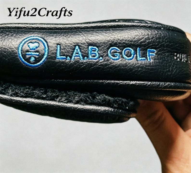 May include: Black golf club head cover with blue embroidered text and logo. The text reads "L.A.B. GOLF". The cover has a textured surface and a soft, fuzzy interior lining. The brand name "Yifu2Crafts" is in the top left corner.