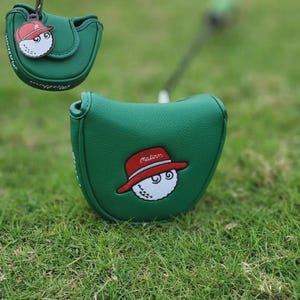 Könnte beinhalten: Grüne Golf-Putter-Schlägerhaube mit einem Cartoon-Golfball, der einen roten Hut trägt. Der Golfball hat zwei Augen und lächelt. Die Haube befindet sich auf einer grünen, grasbewachsenen Oberfläche.