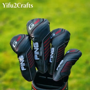 May include: Black golf club head covers with red accents and the brand name "PING" in white. The covers have the text "G410" in white and red. The background is a blurred green.