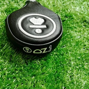 Puede incluir: Funda negra para la cabeza del palo de golf con dise&ntilde;o bordado en blanco. La funda presenta una corona y un dise&ntilde;o circular con el texto "OZ.1". La funda est&aacute; sobre una superficie verde.