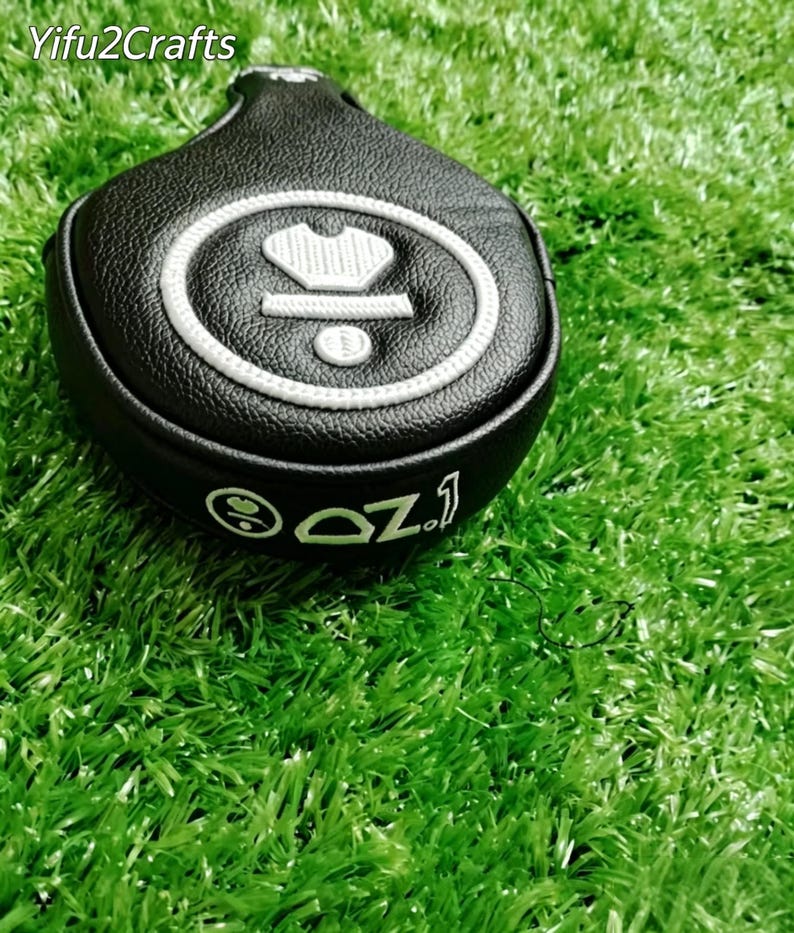 Puede incluir: Funda negra para la cabeza del palo de golf con dise&ntilde;o bordado en blanco. La funda presenta una corona y un dise&ntilde;o circular con el texto "OZ.1". La funda est&aacute; sobre una superficie verde.