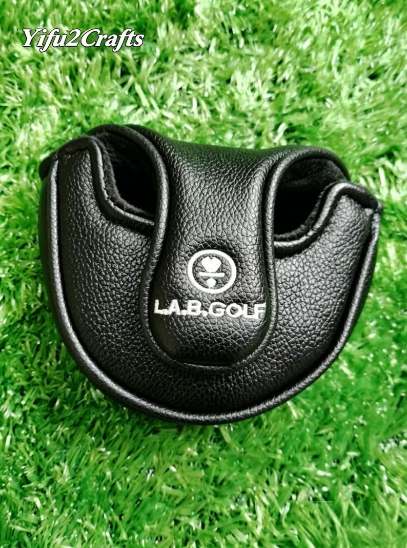 Puede incluir: Funda negra para putter de golf con el texto "L.A.B. GOLF" en blanco. La funda est&aacute; hecha de material texturizado y tiene forma redondeada con dos aberturas. La funda est&aacute; sobre una superficie verde. La marca "Yifu2Crafts" est&aacute; en la esquina superior izquierda.