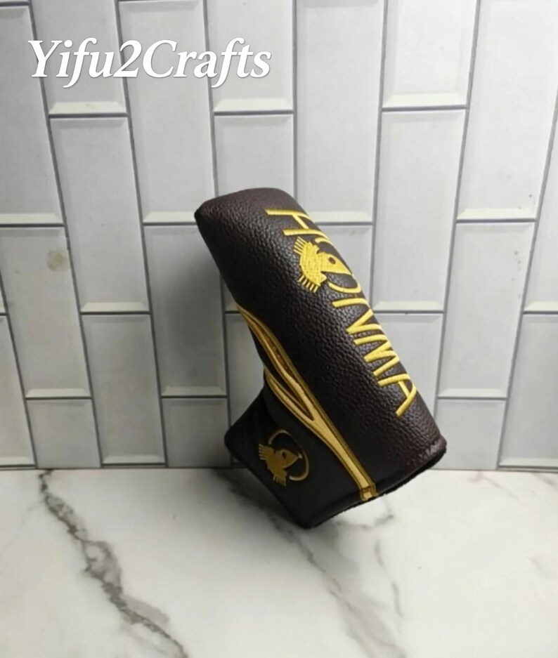 May include: A brown golf club head cover with gold embroidered text and logo. The cover has a textured surface and the text "HONMA" is visible. The logo is a stylized bird. The shop name "Yifu2Crafts" is in the top left corner.