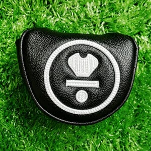 Puede incluir: Funda negra para la cabeza del palo de golf con un dise&ntilde;o bordado en blanco. La funda tiene una superficie texturizada y una forma redondeada. El dise&ntilde;o incluye una corona, una l&iacute;nea horizontal y un c&iacute;rculo. La funda est&aacute; sobre una superficie verde.