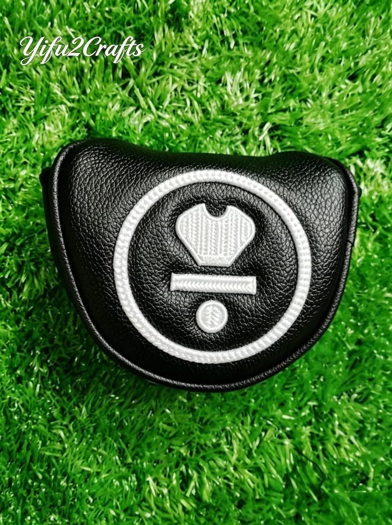 Puede incluir: Funda negra para la cabeza del palo de golf con un dise&ntilde;o bordado en blanco. La funda tiene una superficie texturizada y una forma redondeada. El dise&ntilde;o incluye una corona, una l&iacute;nea horizontal y un c&iacute;rculo. La funda est&aacute; sobre una superficie verde.