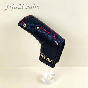 May include: Black golf club head cover with red and gold floral embroidery. The cover has the word "HONMA" in gold lettering. The text "Yifu2Crafts" is in the upper left corner.
