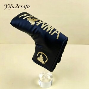May include: A navy blue golf club head cover with gold embroidered text and logo. The cover features the word "HONMA" in gold lettering and a gold bird logo. The cover is displayed on a clear stand.