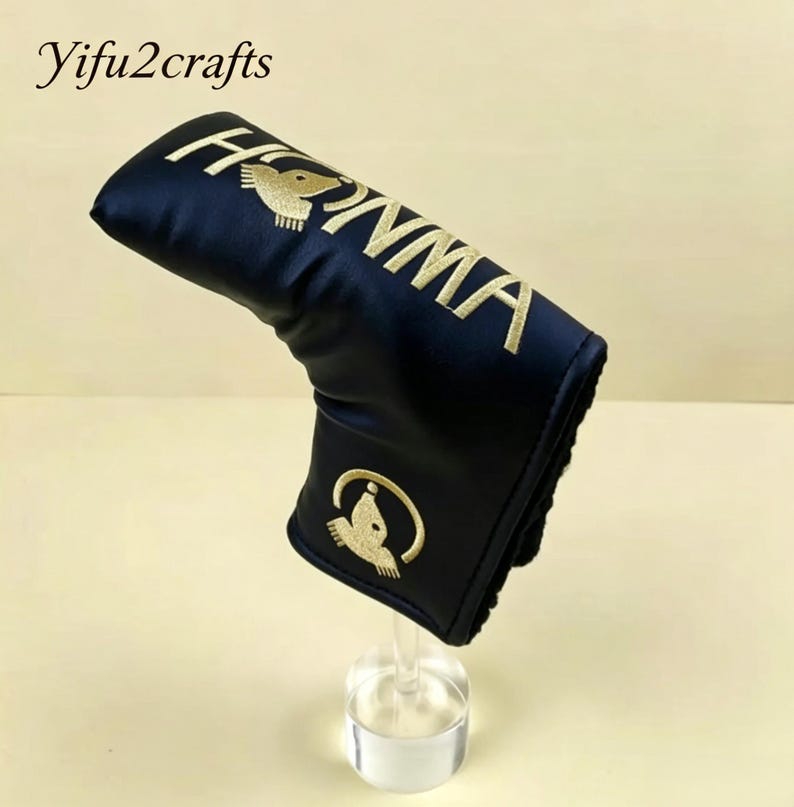 May include: A navy blue golf club head cover with gold embroidered text and logo. The cover features the word "HONMA" in gold lettering and a gold bird logo. The cover is displayed on a clear stand.