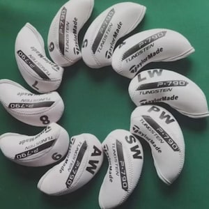 以下が含まれることがあります： 白いゴルフヘッドカバーのセット。各カバーには「P.790 TUNGSTEN TaylorMade」の文字と数字または文字の指定が印刷されています。カバーは緑色の表面に配置されています。